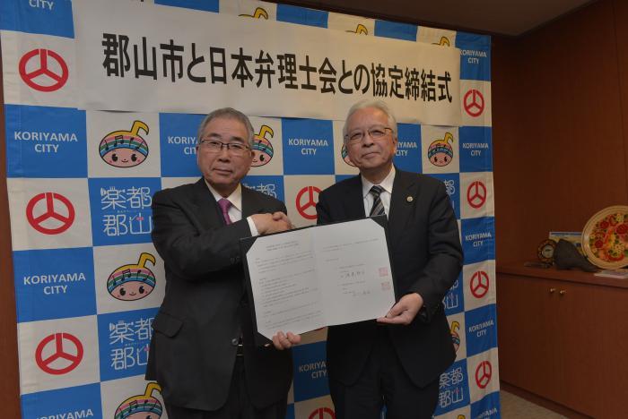郡山市と日本弁理士会との協定締結式にて署名と捺印がされた契約書を持ち握手をする渡邉日本弁理士会会長と品川市長の写真