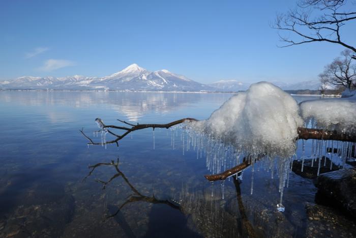 冬の磐梯山と木々の雪解け水がつららになっている猪苗代湖の写真