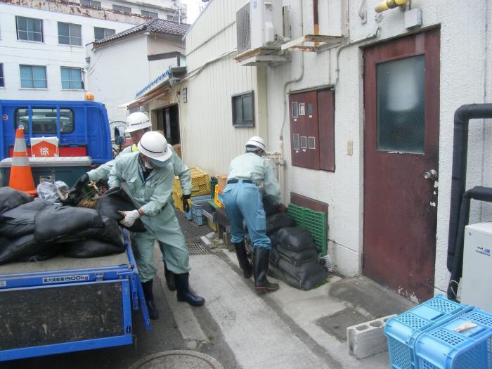 ヘルメットをかぶった3人の男性が、トラックの荷台から建物入り口へと土のうを積んでいる様子