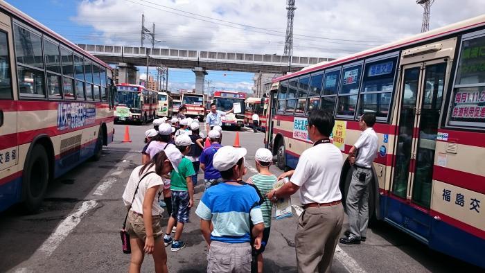 広い敷地内にバスが複数台停車している。それらを見回る小学生の集団と引率する先生方。
